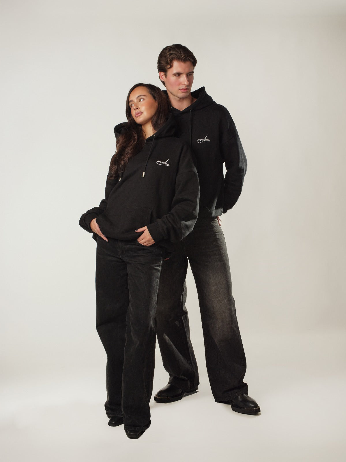 Two models showcase the signature black hoodie from 444 Attire, a new upcoming streetwear clothing brand. The female model stands in front with a relaxed, hands-in-pocket pose, while the male model stands slightly behind her, both dressed in matching black hoodies and loose-fit black jeans. The hoodies have a clean, minimalist design with a small white 444 Attire logo printed on the left chest, emphasizing subtle branding and modern aesthetics. Shot in a bright studio on a light, neutral background, the image highlights the unisex, oversized fit and versatile style of the hoodie, perfect for couples, casual everyday wear, and contemporary urban fashion.