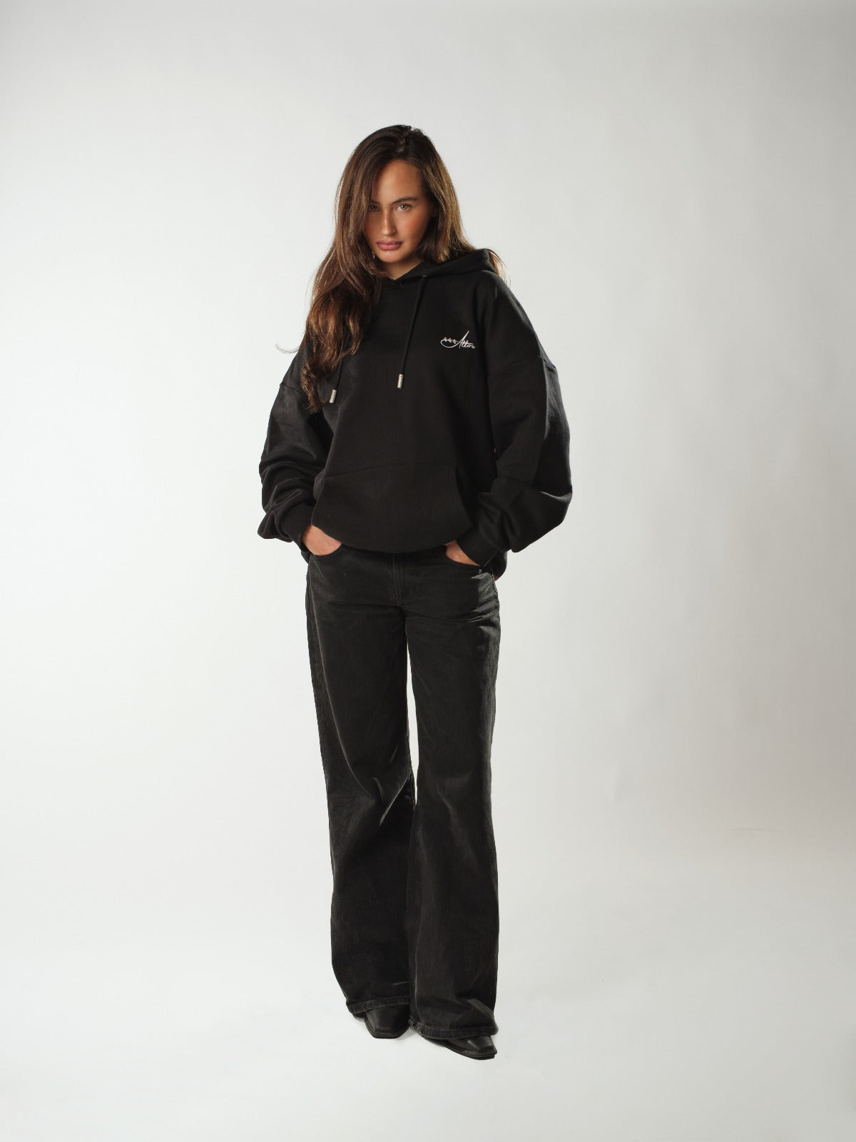 Female model wearing the signature black hoodie from 444 Attire, a new upcoming streetwear clothing brand. She stands in a bright studio with a neutral background, hands in the kangaroo pocket, styled with loose-fit black jeans. The hoodie features a clean, minimalist design with a small white 444 Attire logo on the left chest, highlighting its oversized, comfy unisex fit. This image showcases a modern women’s black hoodie outfit perfect for everyday casual and urban streetwear looks.
